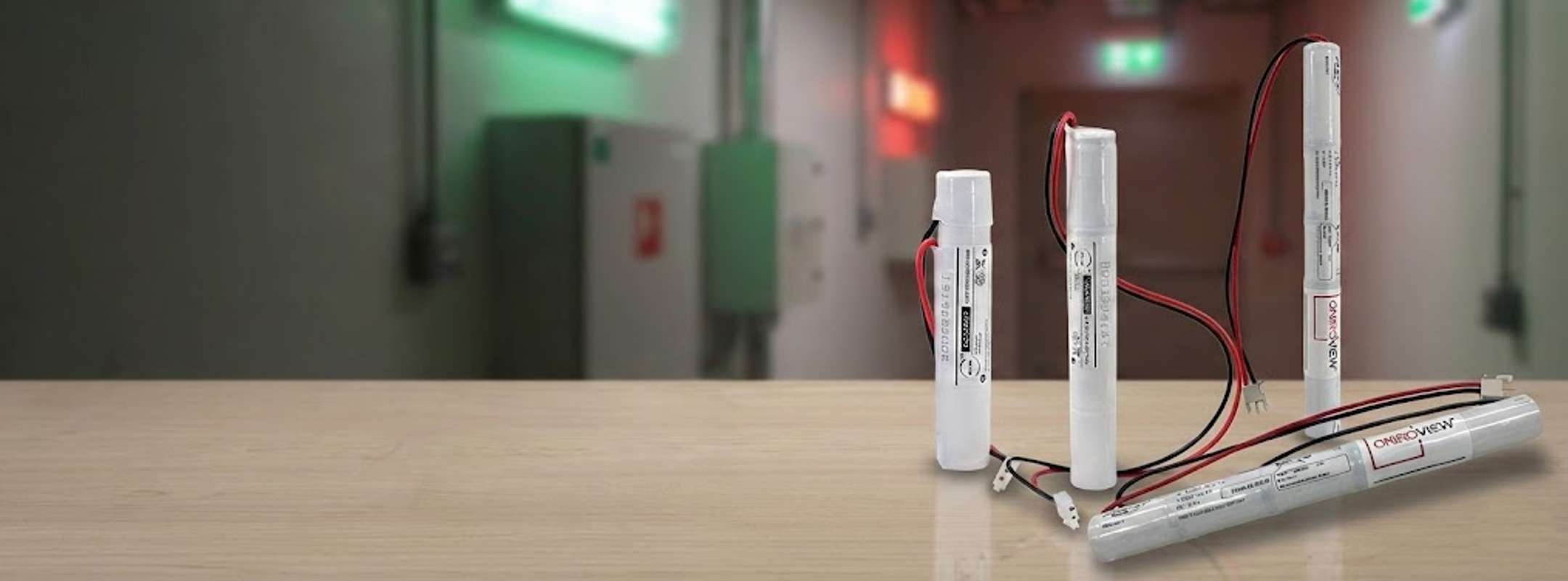 Primo piano di una selezione di pacchi batteria ricaricabili bianchi Oniroview per lampade di emergenza, di diverse dimensioni con cavi rossi e neri. Sono appoggiati su un banco di legno, con uno sfondo sfocato di un corridoio con luci di sicurezza verdi e rosse accese.